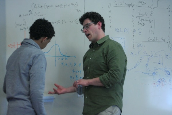 Standing in front of a whiteboard with equations, a male Mitsubishi Electric engineer mentors a young man.