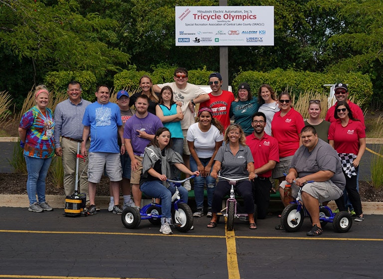 Twenty Mitsubishi Electric volunteers in a parking lot, 3 on large tricycles, pose in front of a 'Tricycle Olympics' sign