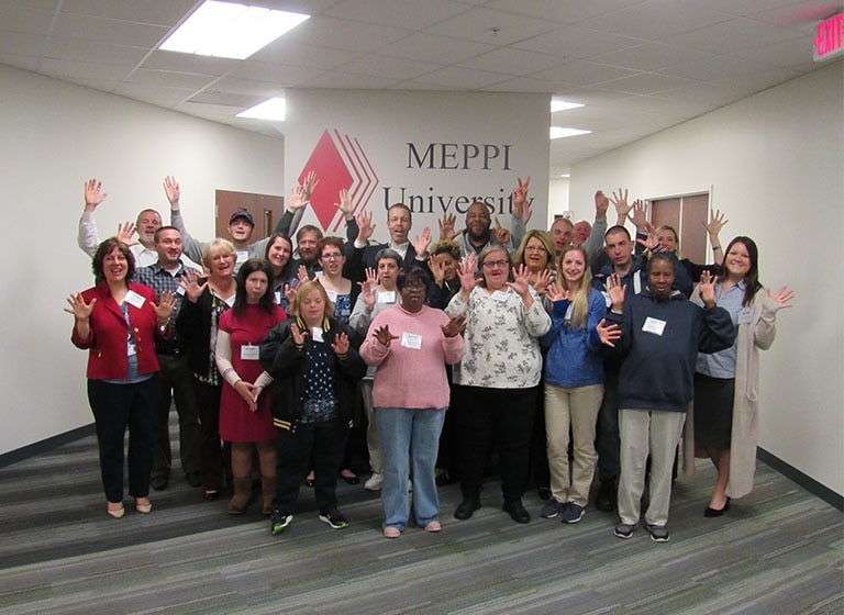 Twenty-three Mitsubishi Electric volunteers and clients from Life'sWork stand in front of a 'MEPPI University' sign.