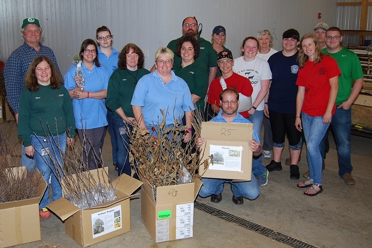 Seventeen Mitsubishi Electric volunteers pose hold boxes containing saplings in a barn.
