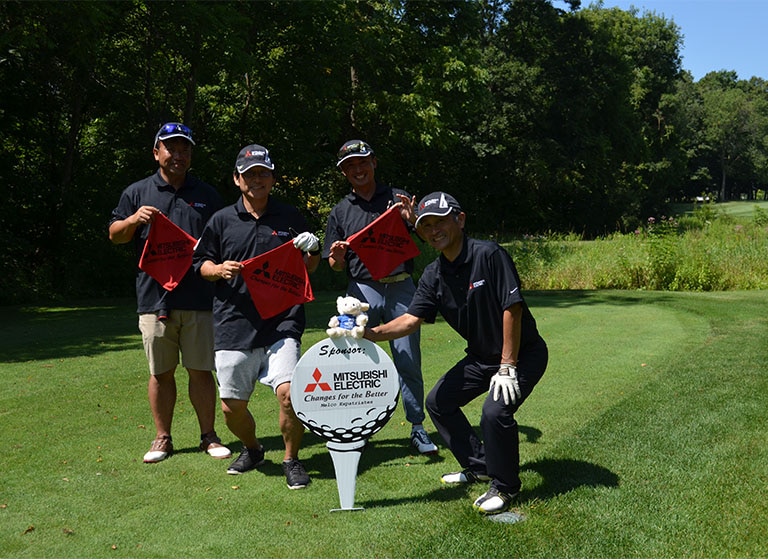 Four Japanese men in golf outfits stand in front of a hole marker that reads 'Mitsubishi Electric.'